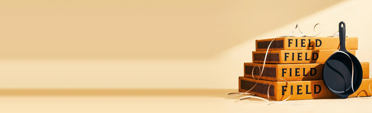 A stack of Field skillet boxes with a black cast iron skillet leaning against them, ribbon curling around, on a warm cream background